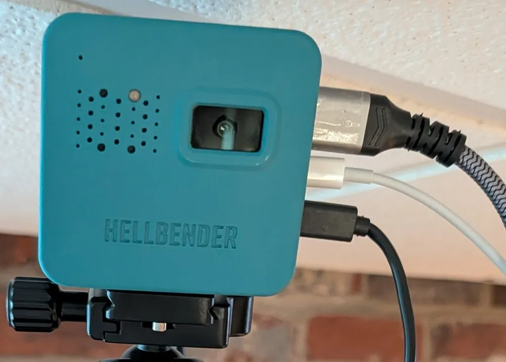 The RoomState sensor in place: a teal Hellbender Tendryl camera clamped to a tripod head against a brick wall, close under the ceiling, with power and data cables running from its side.